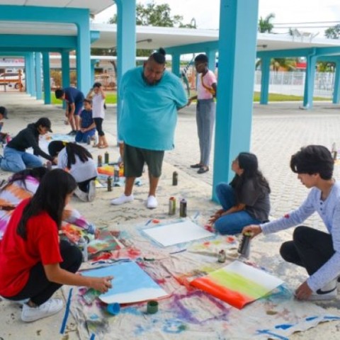 Students paint outside under a walkway while a teaching artist oversees their work.