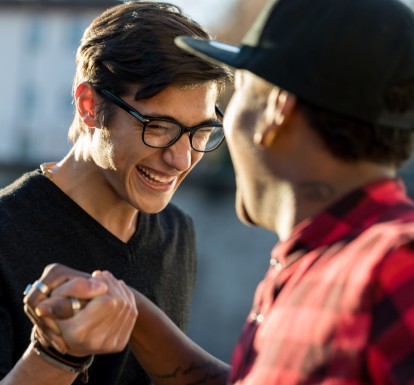 A young man and a mentor grab hands and smile at each other outside.