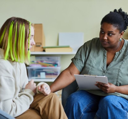 A serious-looking teenager with neon hair extensions talking with a school counselor, who is comforting the student by placing her hand on the student's rm.