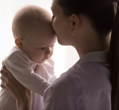 A mother holds her baby with her hands and kisses the baby on the forehead, against a white background.