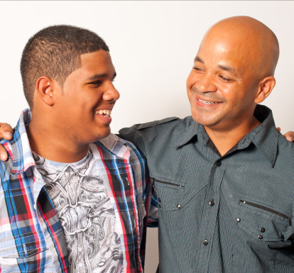 A father and teenage son smile at each other, with arms around each other. 