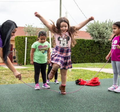 Three little girls play jump rope with a child care teacher on a playground.