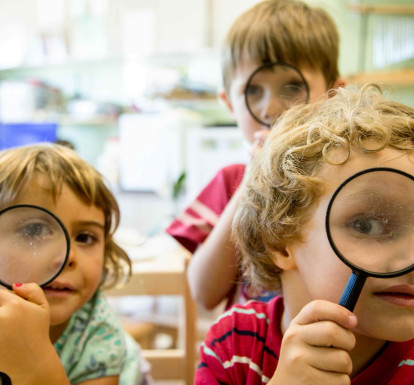 Three children in a preschool program look at the camera through magnifying lenses.