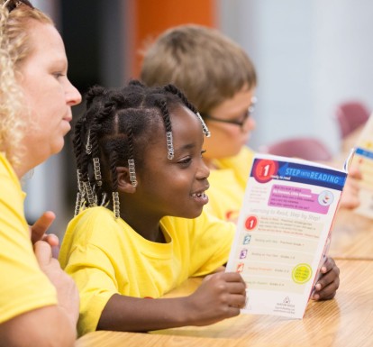 A teacher watches as two children read books at summer camp.