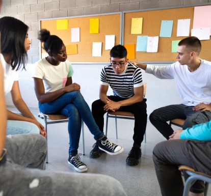 Teens sit in a circle talking and comforting one young man who looks upset.