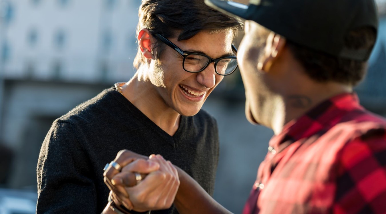 A young man and a mentor grab hands and smile at each other outside.