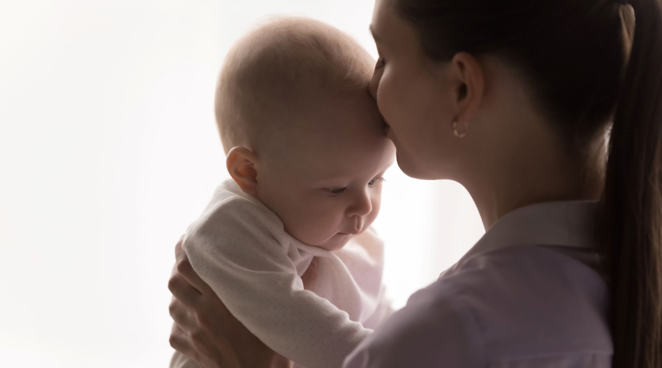 A mother holds her baby with her hands and kisses the baby on the forehead, against a white background.