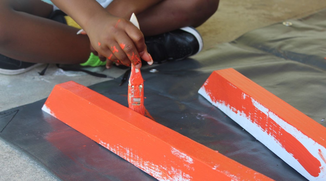 A child sits cross-legged on the floor using orange paint to paint the legs of a picnic table. 