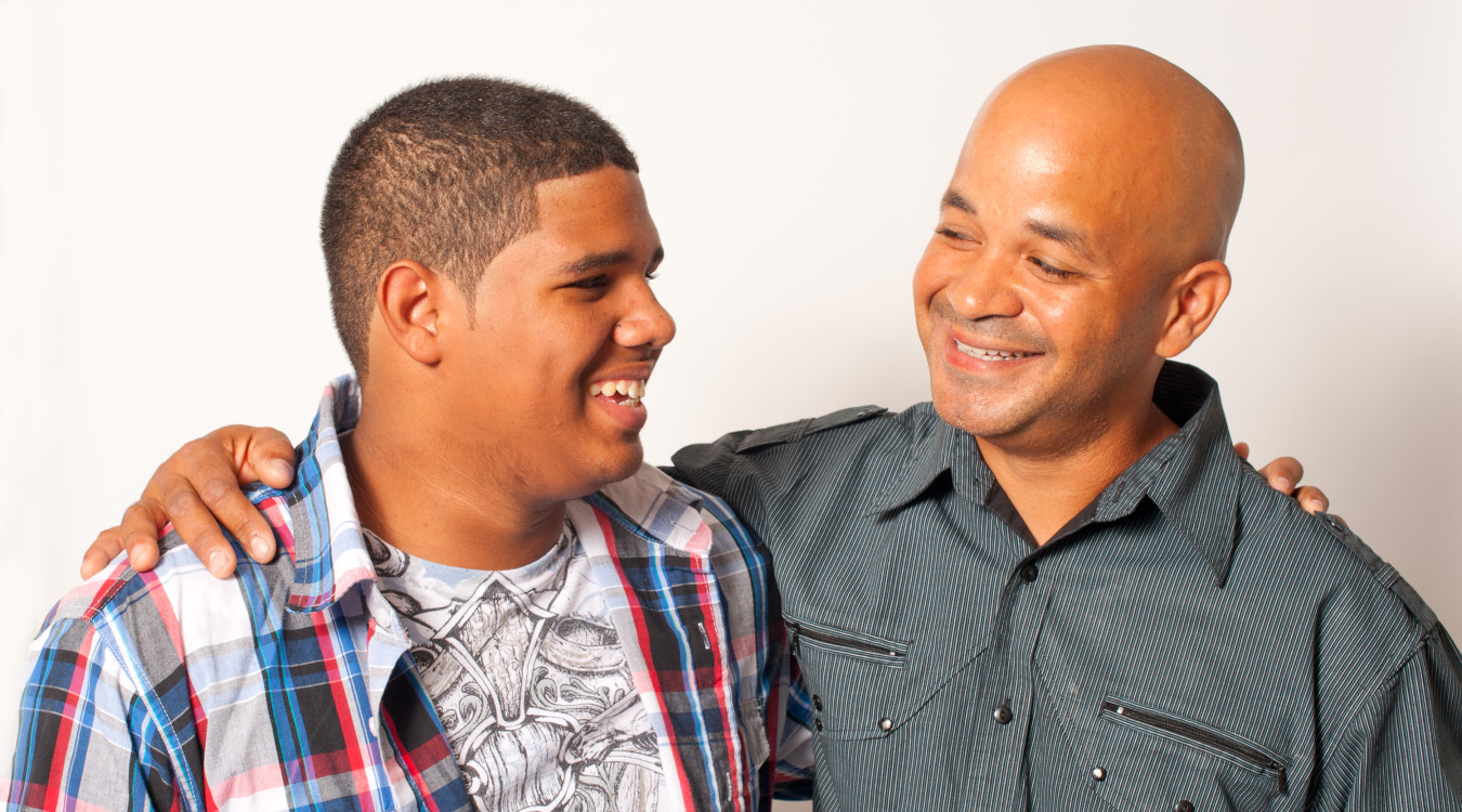 A father and teenage son smile at each other, with arms around each other. 