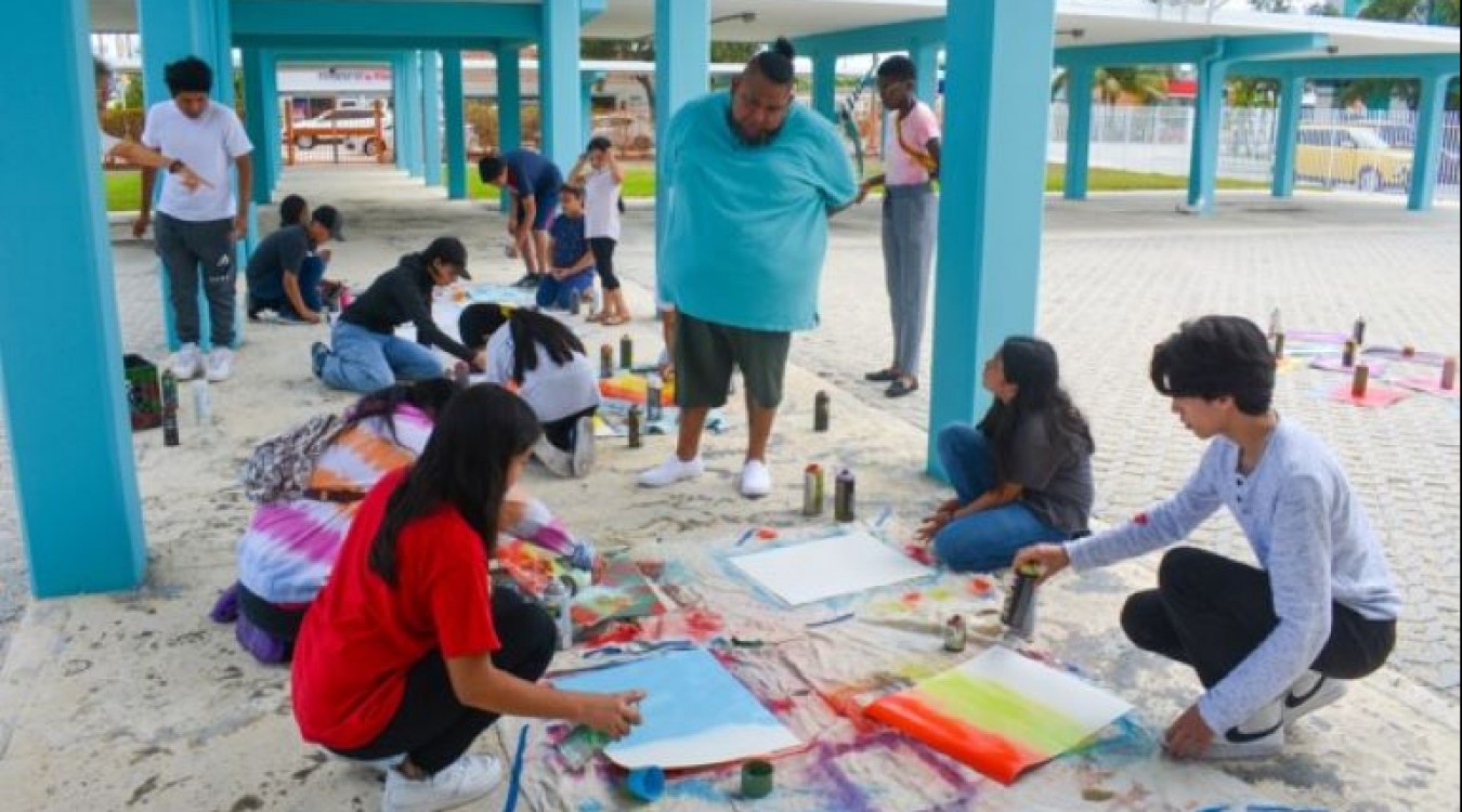Students paint outside under a walkway while a teaching artist oversees their work.