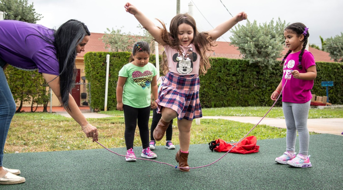 Three little girls play jump rope with a child care teacher on a playground.