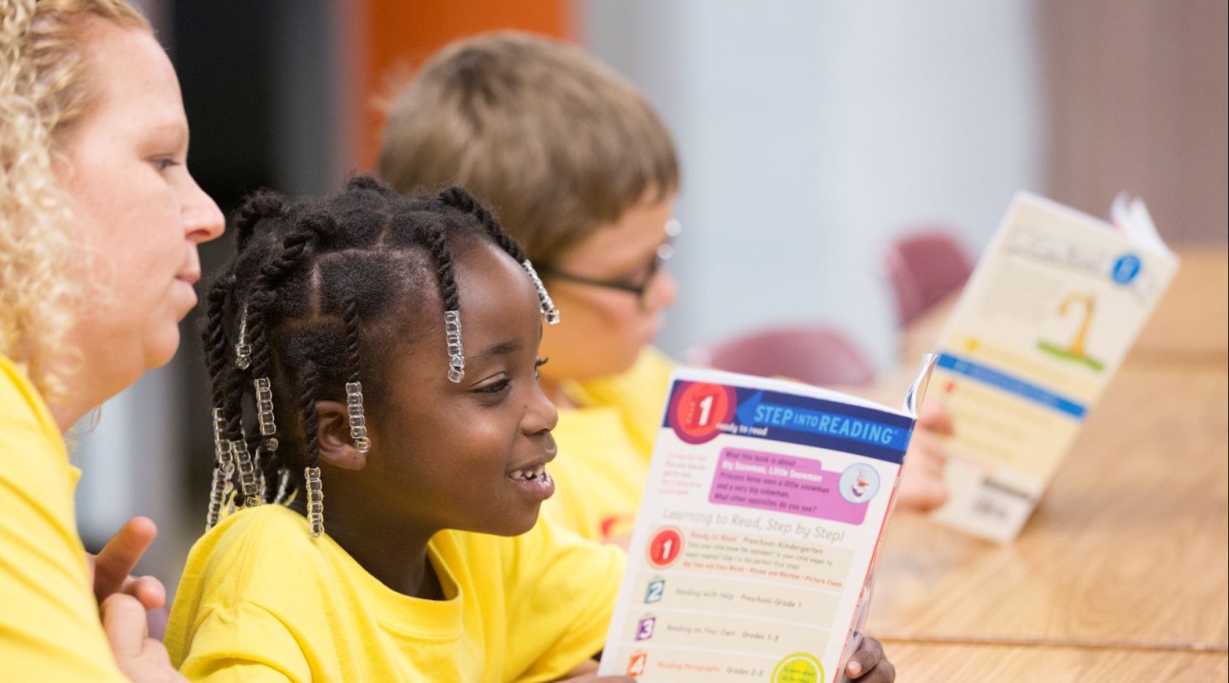 A teacher watches as two children read books at summer camp.