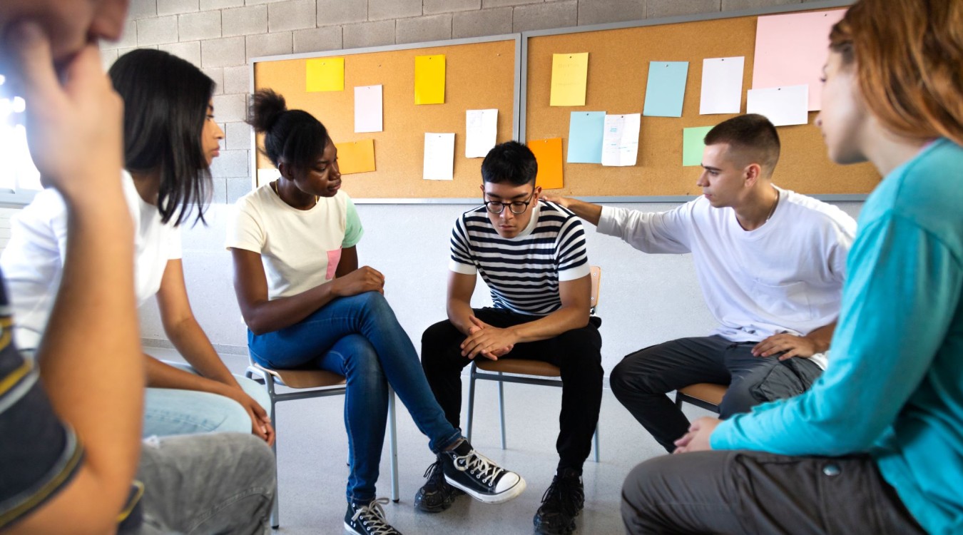 Teens sit in a circle talking and comforting one young man who looks upset.