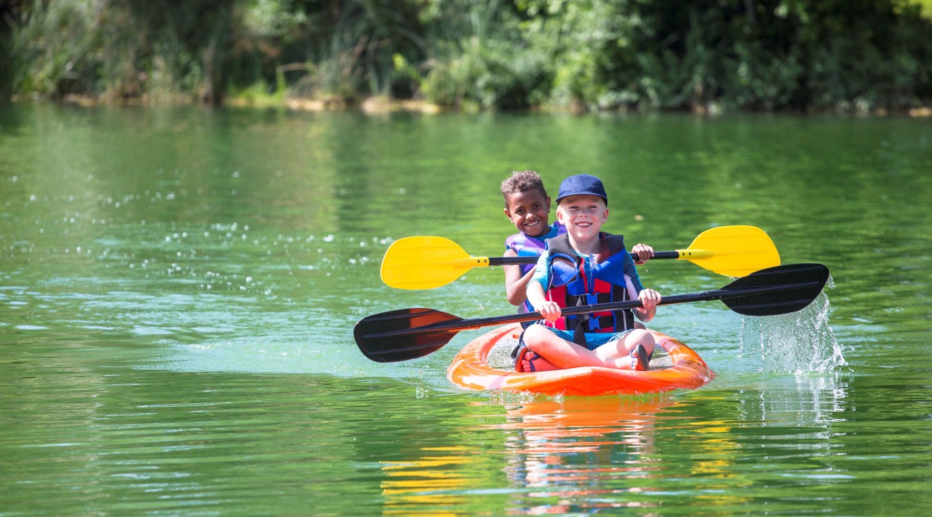 Two boys wearing life jackets in a kayak smiling while rowing through a river.