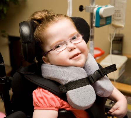A girl with special needs strapped in to a wheelchair with a special cushion to hold her head up. 