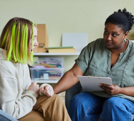 A serious-looking teenager with neon hair extensions talking with a school counselor, who is comforting the student by placing her hand on the student's rm.