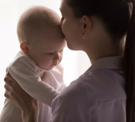 A mother holds her baby with her hands and kisses the baby on the forehead, against a white background.