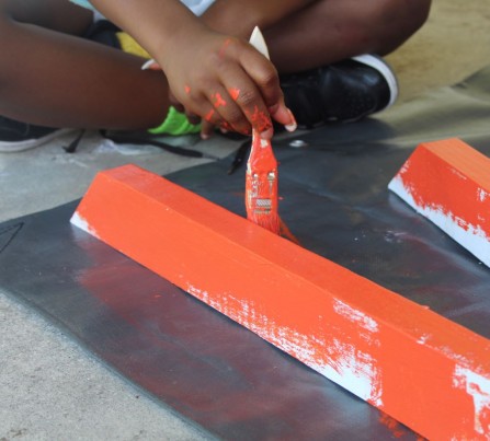 A child sits cross-legged on the floor using orange paint to paint the legs of a picnic table. 