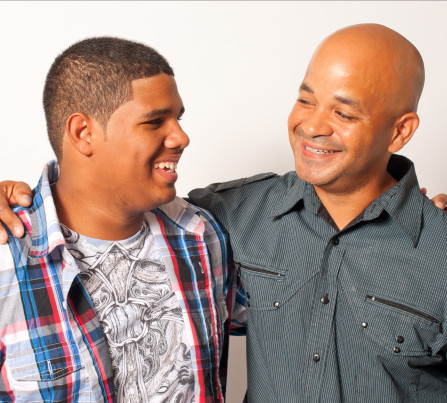A father and teenage son smile at each other, with arms around each other. 