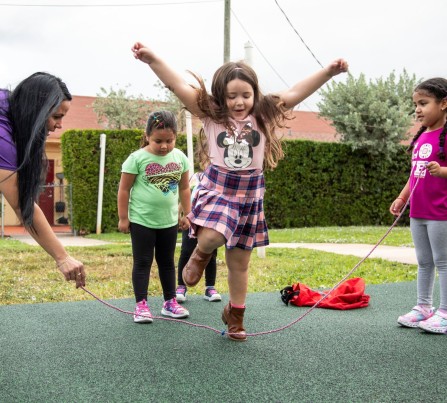 Three little girls play jump rope with a child care teacher on a playground.