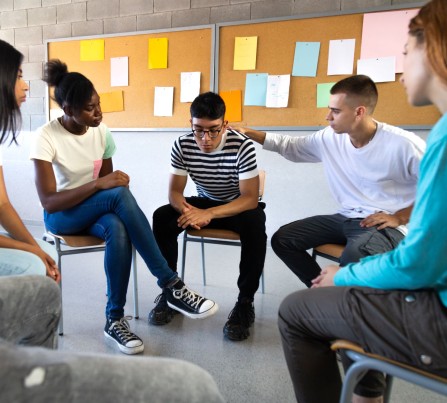 Teens sit in a circle talking and comforting one young man who looks upset.