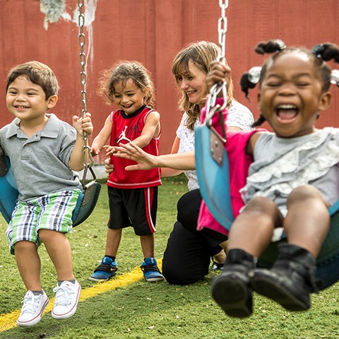 Two happy young children play on swings, while a preschool teacher shows another child how to push the swings.