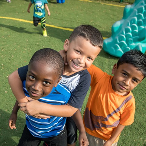 Three preschool boys with their arms around each other, smiling, while on a playground.