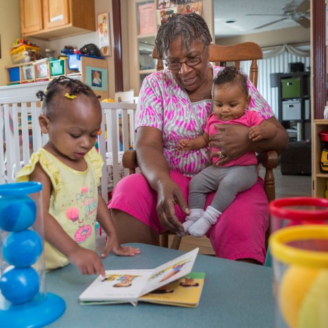 A child care provider holds a baby on her lap while reading a book with a toddler.