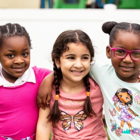 Three smiling little girls with their arms around each other sit outside while at child care. 