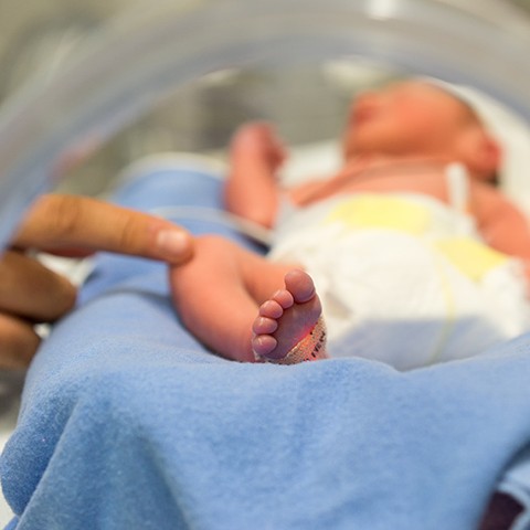 Premature baby in an incubator with a man's finger touching the baby's knee.