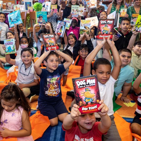 A classroom full of children sitting on mats holding up books and smiling.