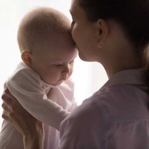A mother holds her baby with her hands and kisses the baby on the forehead, against a white background.