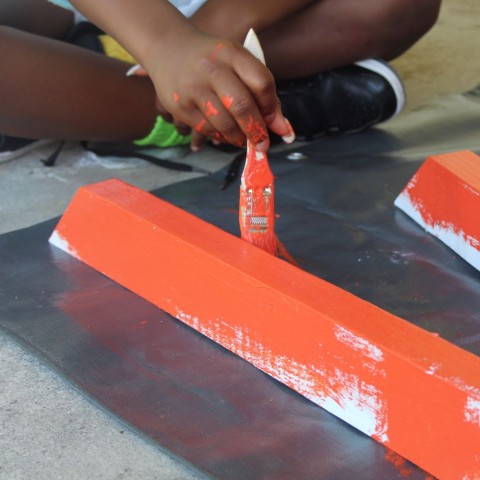 A child sits cross-legged on the floor using orange paint to paint the legs of a picnic table. 