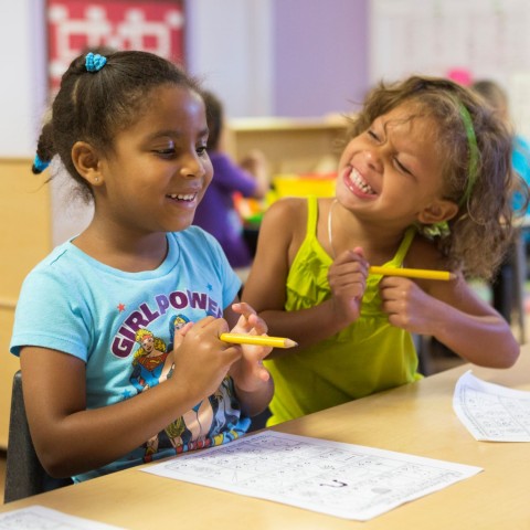 Two smiling little girls sit at a desk with pencils and paper.