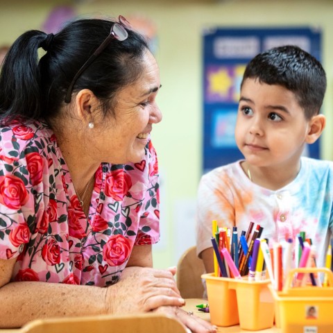 A child care teacher leans over a table and smiles at a little boy, who has art supplies in front of him.