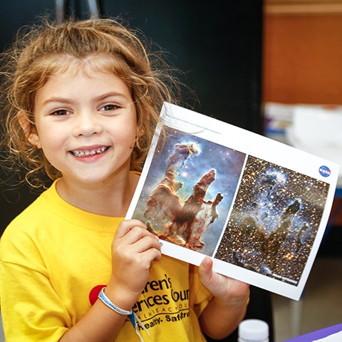 Young girl, smiling, holds up pictures of stars and space during a summer learning class.
