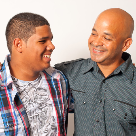 A father and teenage son smile at each other, with arms around each other. 
