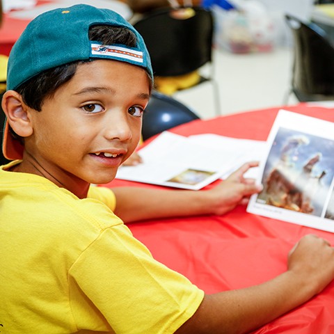 Boy in a CSC T-shirt sits at a table reading a book during a summer camp program.