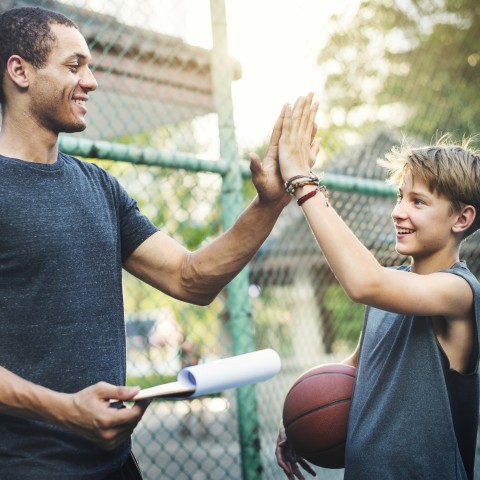 Man, who's holding a clipboard, high-fiving a pre-teen, who's holding a basketball. Both are smiling.
