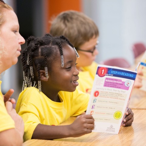 A teacher watches as two children read books at summer camp.