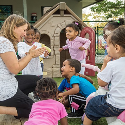 Child care teacher claps hands and smiles while playing with preschoolers outside (photo taken pre-COVID).