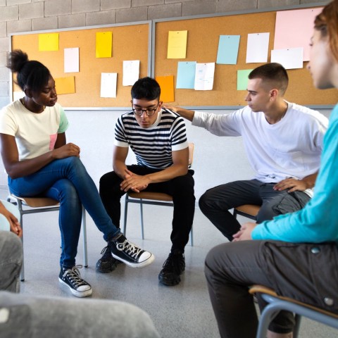 Teens sit in a circle talking and comforting one young man who looks upset.