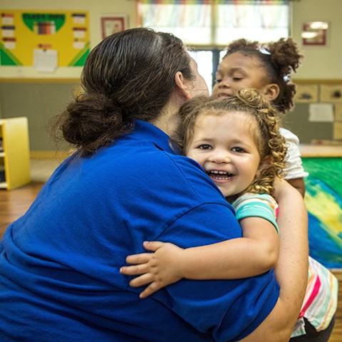 Smiling little girl hugging child care teacher in a preschool classroom.