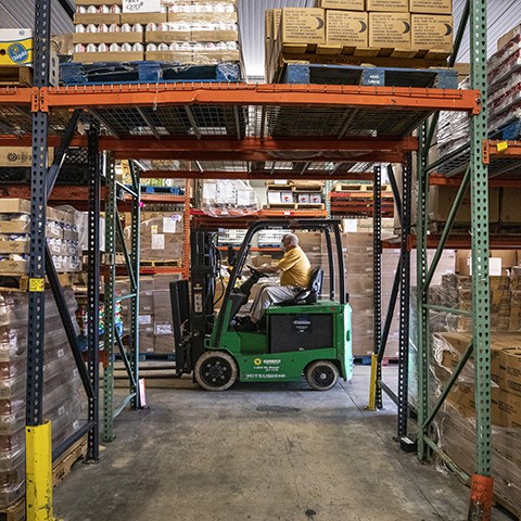 Man on a forklift in the Palm Beach County Food Bank's warehouse, which has shelves filled with boxes and cans of food