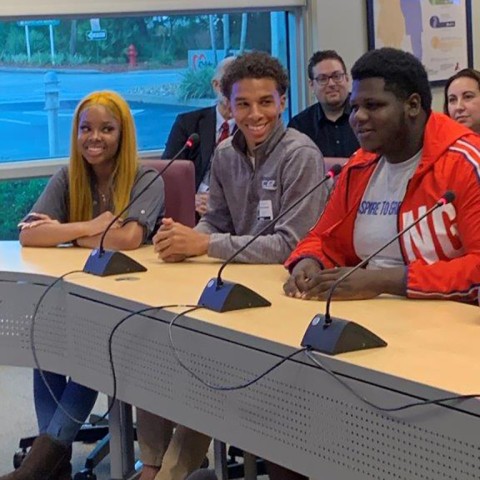 Three high school students from the Student Aces program smile while they sit at a conference table presenting their grant-making project to Children's Services Council's board. 