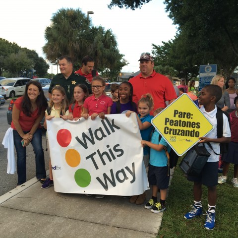 Children and adults gather together before walking to school for International Walk to School Day. The elementary school-age children hold a sign that says "Walk this Way."