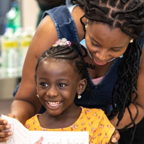 A little girl presents her booklet about a red bird while her mom stands proudly behind her helping her hold it up.